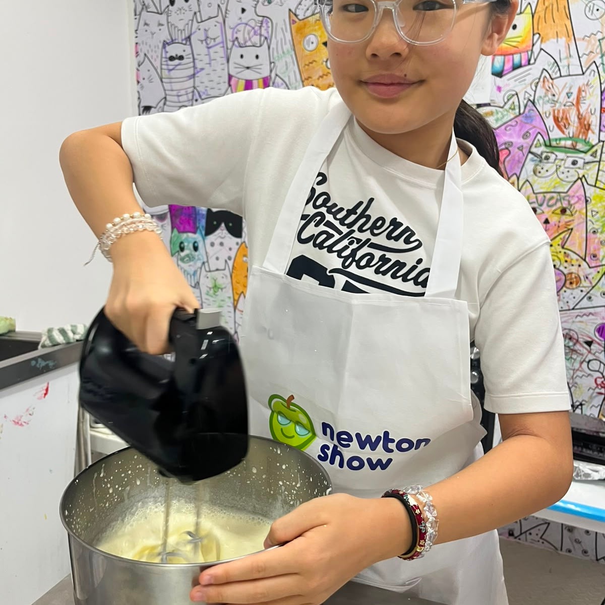 Girl in a Newtonshow apron using a hand mixer during a camp cooking activity